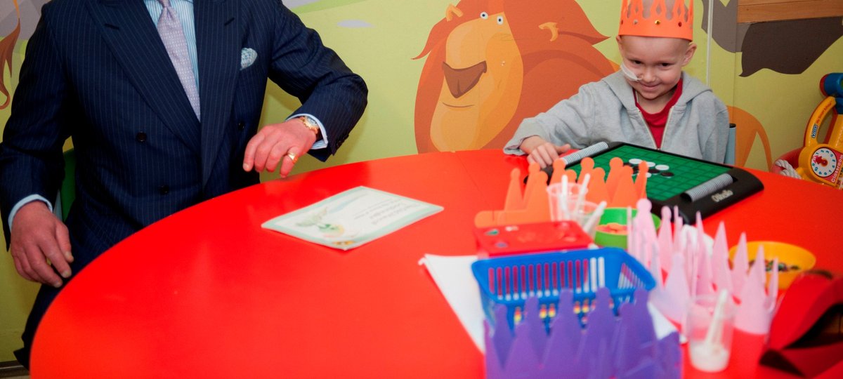King Charles sitting at a round red table in a black suit next to a patient in a orange crown playing a game. They are sat in front of a wall with cartoon lions.