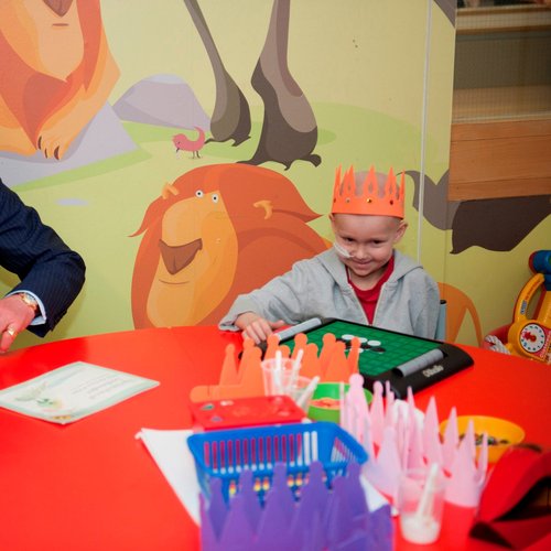 King Charles sitting at a round red table in a black suit next to a patient in a orange crown playing a game. They are sat in front of a wall with cartoon lions.