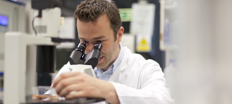Man in white lab coat looking through a microscope in a research lab