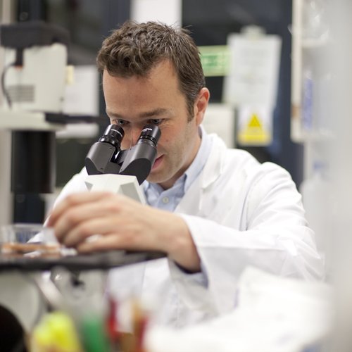Man in white lab coat looking through a microscope in a research lab