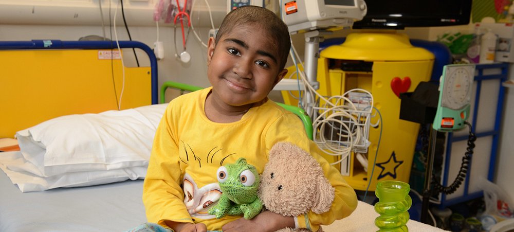 Child, Keano, smiling and sitting up on hospital bed holding stuffed teddy and lizard. There is a game next to him on the bed.