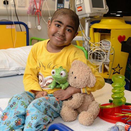 Child, Keano, smiling and sitting up on hospital bed holding stuffed teddy and lizard. There is a game next to him on the bed.