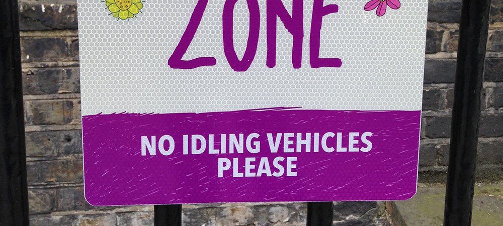 Photo of a sign reading "Clear air zone" and decorated with children's drawings of flowers and butterflies. The caption beneath reads "No idling vehicles please".