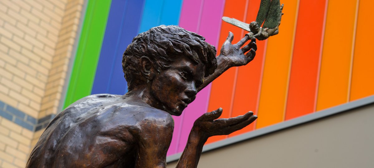 Photo of Peter Pan statue with his left hand stretched up to the sky. On his index finger is a thimble, which his trusty friend Tinkerbell is trying to wrestle off. In Peter’s other hand he has a palm of invisible fairy dust which he is blowing onto passers-by.