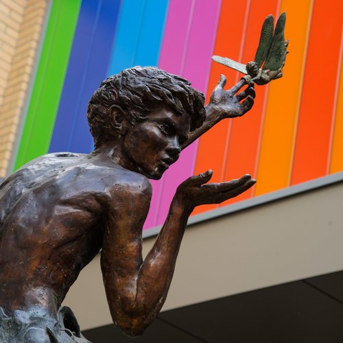 Photo of Peter Pan statue with his left hand stretched up to the sky. On his index finger is a thimble, which his trusty friend Tinkerbell is trying to wrestle off. In Peter’s other hand he has a palm of invisible fairy dust which he is blowing onto passers-by.