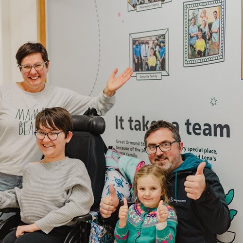 Amelia and Lena and their parents stand in front of the Family Wall smiling.
