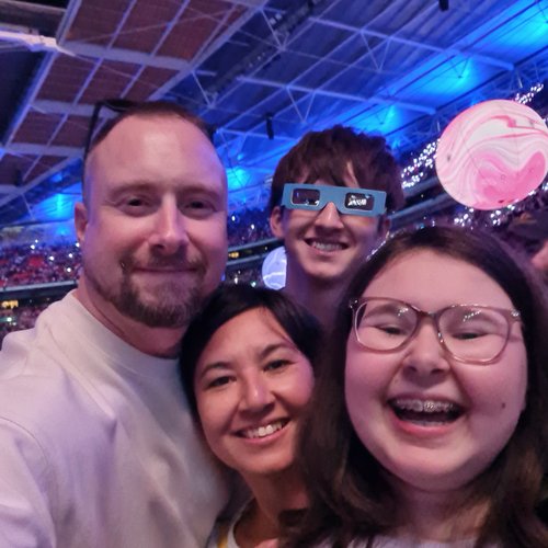 A family smile at the camera with a stadium roof in the background. They are all smiling and laughing and the boy at the back is wearing novelty glasses