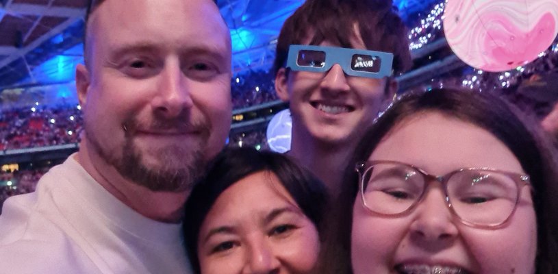 A family smile at the camera with a stadium roof in the background. They are all smiling and laughing and the boy at the back is wearing novelty glasses