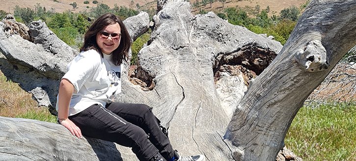 A young woman sits on a fallen tree and smiles at the camera