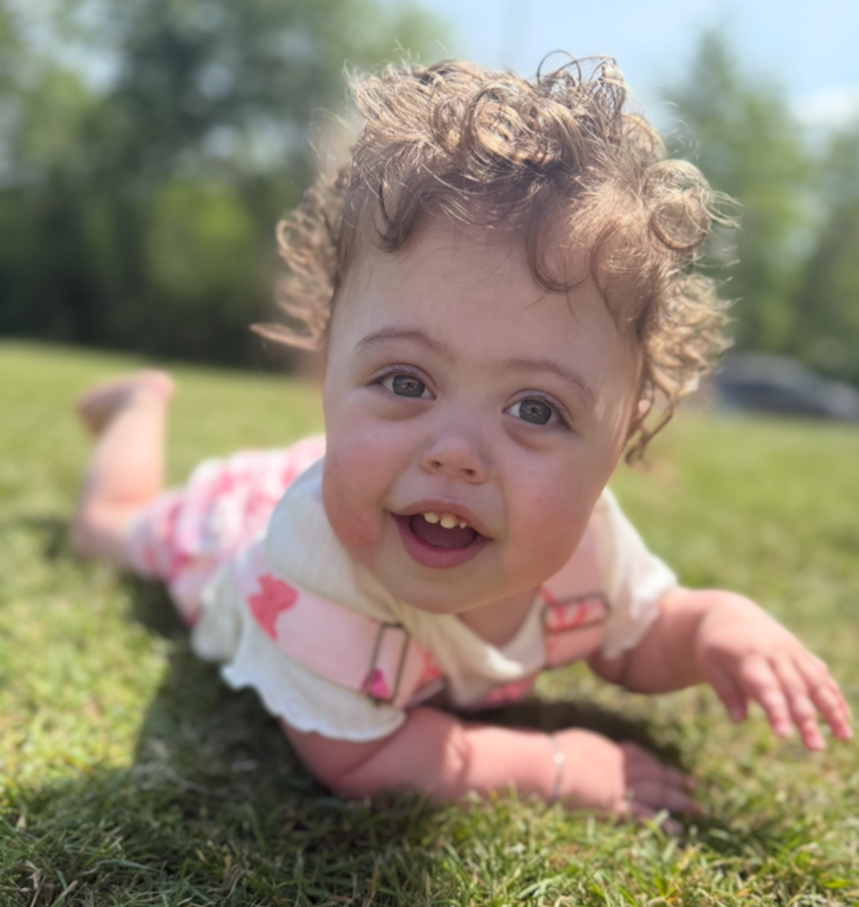 A baby girl with curly hair lies on the grass and smiles at the camera