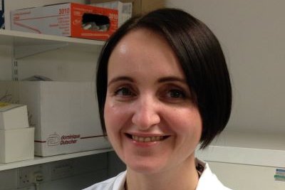 Amy McTague stands in a black t-shirt and white lab coat. She has her arms crossed and is smiling at the camera, she has a short dark brown bob.