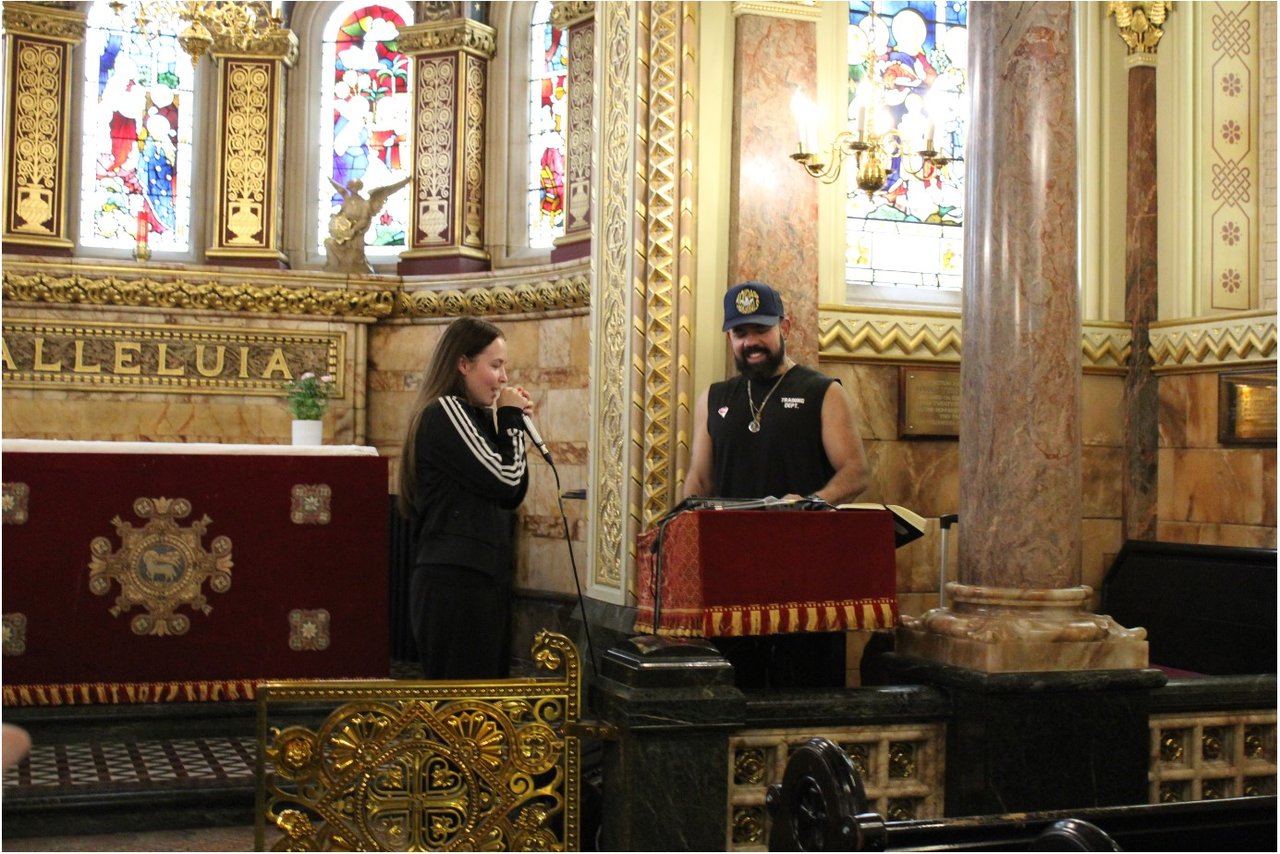 A young person and a man in a cap. One is holding a microphone. They are in a Chapel, surrounded by stained glass windows.