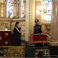 A young person and a man in a cap. One is holding a microphone. They are in a Chapel, surrounded by stained glass windows.