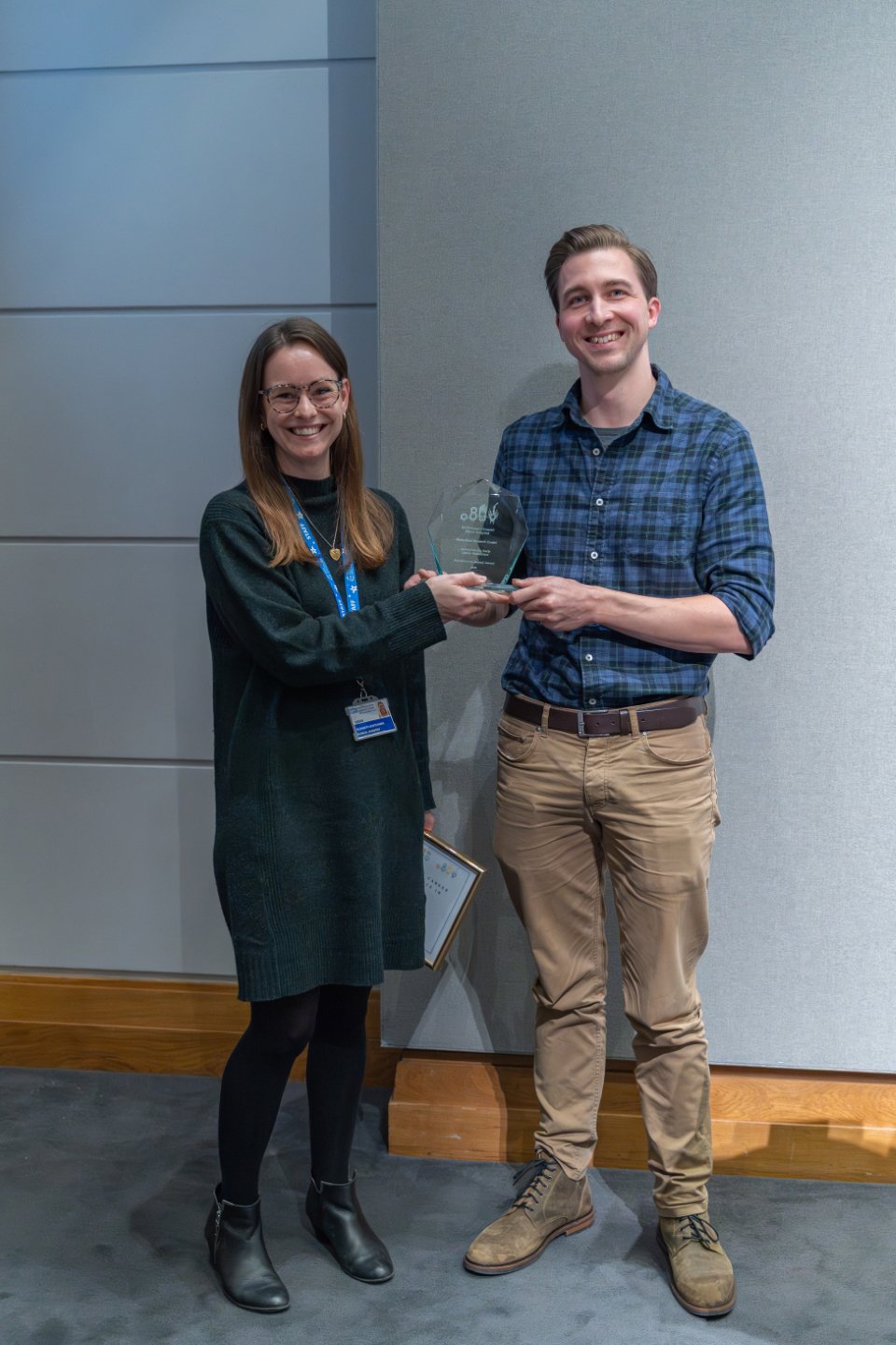 A man in a check blue shirt smiles at the camera as he receives an award from a smiling woman in a black long sleeve dress