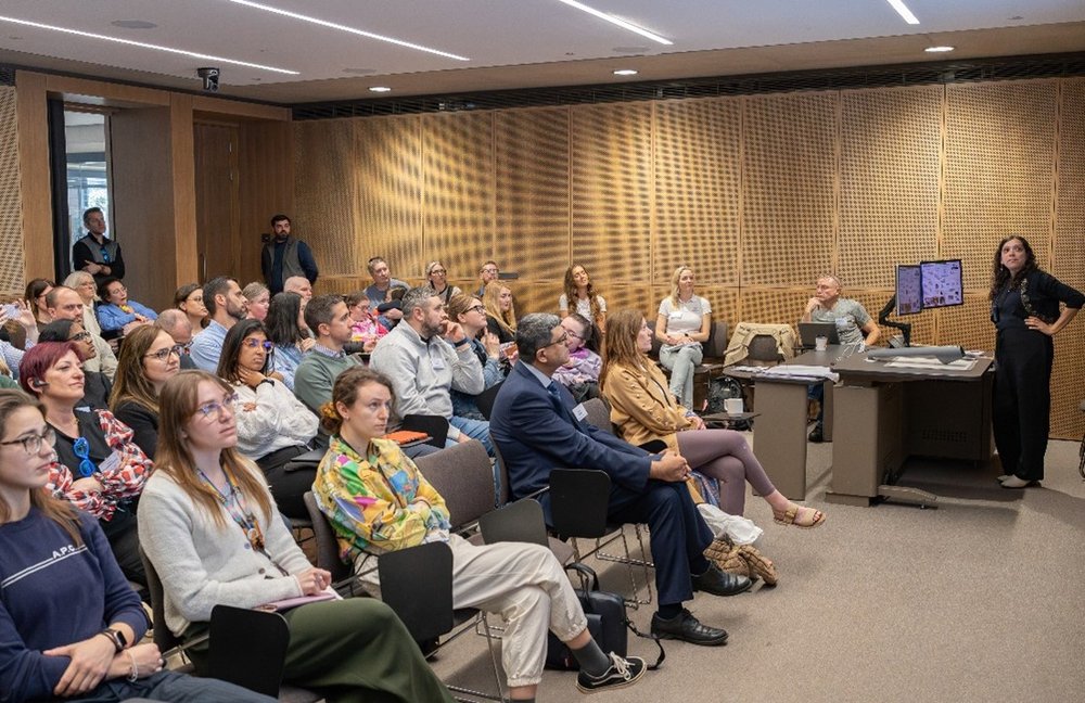 A room full of people look at a screen (out of sight), while a woman stands and gestures at a speaker's position