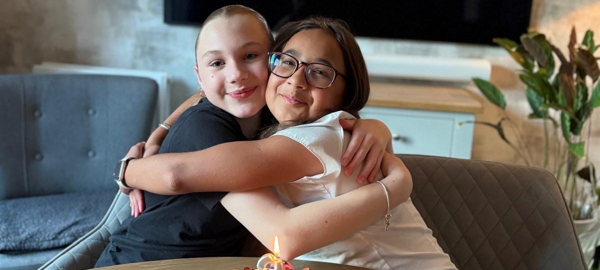 Two young people sat on chairs smiling and hugging. In front of them is a cake with a number 2 candle on it. Behind them is a screen that says 'Happy Anniversary'.