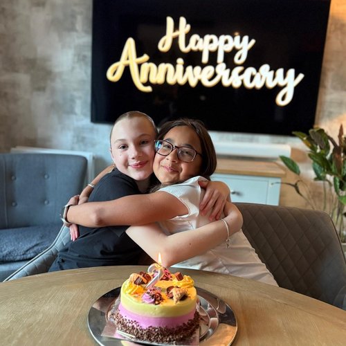 Two young people sat on chairs smiling and hugging. In front of them is a cake with a number 2 candle on it. Behind them is a screen that says 'Happy Anniversary'.