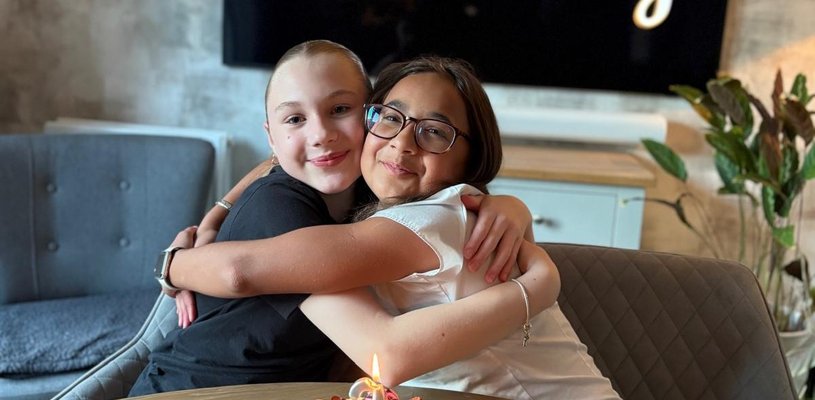 Two young people sat on chairs smiling and hugging. In front of them is a cake with a number 2 candle on it. Behind them is a screen that says 'Happy Anniversary'.