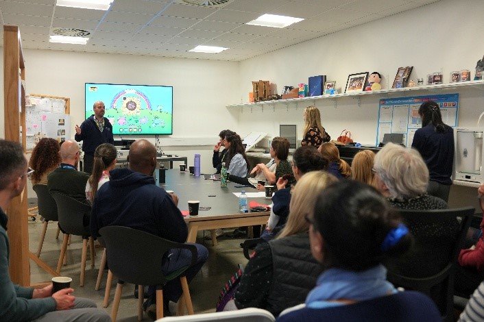 A group of 10-15 people are gathered around a table, attentively watching someone give a presentation at the front of the room