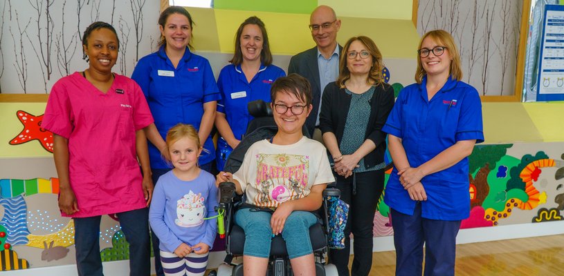 A lady in a wheelchair wearing. She is surrounded by six men and women, and a young girl who wearing a blue top.