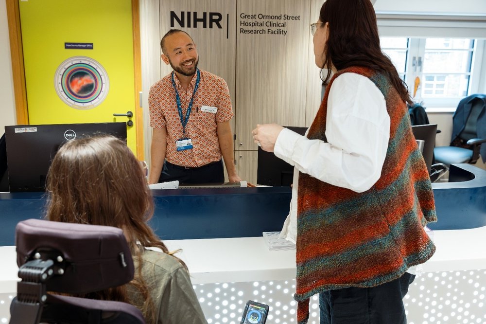 A smiling man is stood behind a reception desk with the sign 'NIHR GOSH Clinical Research Facility' and you can see the back of a young woman in a wheelchair and a woman standing up approaching him.
