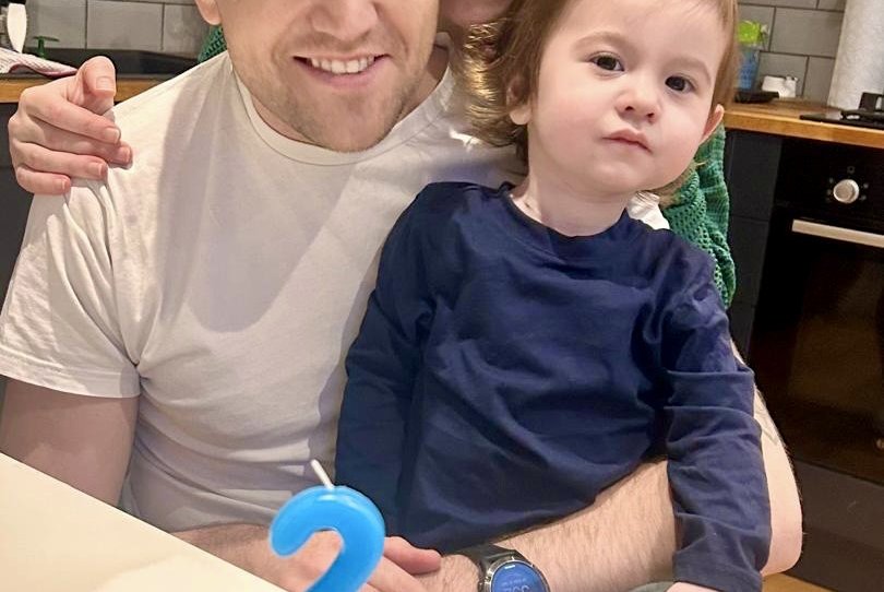 A family scene of a man, woman and young boy smiling widely in a kitchen, cuddled around a cake with a big 2-shaped blue candle.