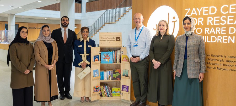 A group of seven people standing in a modern, well-lit lobby beside a bookshelf filled with children's books. They are smiling, projecting a sense of achievement and collaboration. The wooden wall behind them displays the "Zayed Centre for Research into Rare Disease in Children" sign. All are dressed in professional attire.