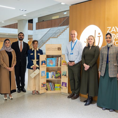 A group of seven people standing in a modern, well-lit lobby beside a bookshelf filled with children's books. They are smiling, projecting a sense of achievement and collaboration. The wooden wall behind them displays the "Zayed Centre for Research into Rare Disease in Children" sign. All are dressed in professional attire.