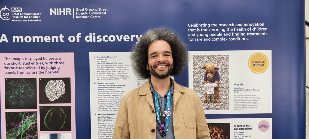 A man stands with his hands in his pockets, smiling broadly. He stands in front of a display bord with lots of images on it