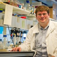 A man in a white lab coat is seated at a lab bench and smiles at the camera. He is wearing glasses