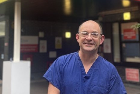 A man in glasses and blue scrubs smiles at a camera in front of a colourful background