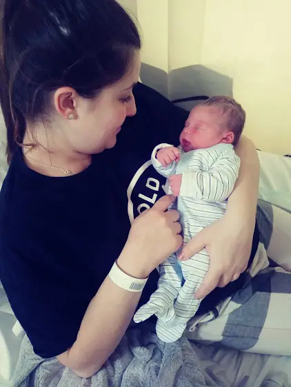 A mum sits on a hospital bed holding a newborn baby dressed in a pale, striped sleepsuit. The mum wears a dark top and a hospital wristband, gently supporting the baby in both arms. The baby is lying on their back with eyes closed, while the mum looks down toward the baby. Pale walls and bedding are visible in the background, showing a hospital bedroom.