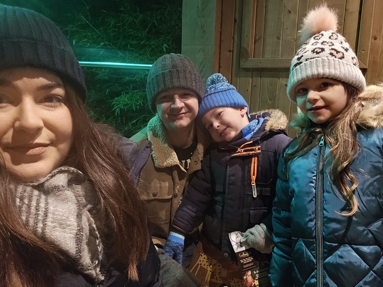 Four people pose closely together for a selfie outdoors at night, wearing winter coats, hats, and scarves. A wooden fence and structure form the background, with green lighting illuminating part of the scene. One person on the right wears a white hat with a pom‑pom, another in the centre wears a grey beanie, and a child leans in between them wearing a blue hat and gloves.