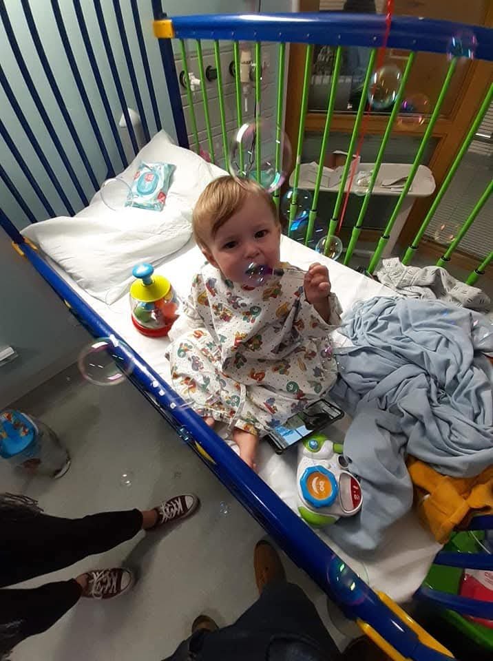 A young child sits upright in a hospital crib with blue and green rails, playing with bubbles. The child wears a patterned hospital gown and holds a bubble wand near their mouth. The crib contains a pillow, blanket, wipes, and several toys, including a colourful rattle and a small electronic toy. A smartphone rests beside the child on the mattress. The floor below is visible with an adult’s shoes partly in frame, and medical equipment and cabinets can be seen in the background of the hospital room.