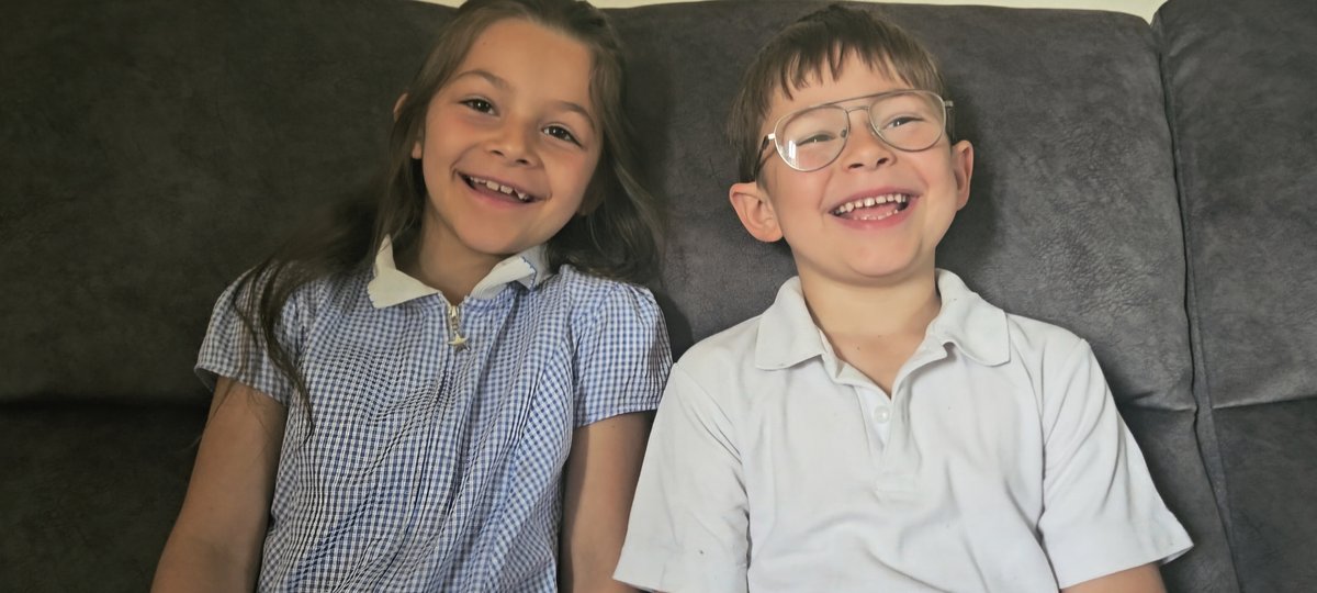 Two children sit side by side on a dark sofa, facing the camera. One child wears a blue-and-white gingham school uniform dress with a white collar, while the other wears a light-colored polo shirt and glasses. Both are smiling broadly, showing missing or growing teeth, with long hair on one child and short hair on the other. The background is a plain wall above the sofa, with soft, even indoor lighting.