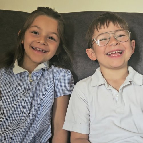 Two children sit side by side on a dark sofa, facing the camera. One child wears a blue-and-white gingham school uniform dress with a white collar, while the other wears a light-colored polo shirt and glasses. Both are smiling broadly, showing missing or growing teeth, with long hair on one child and short hair on the other. The background is a plain wall above the sofa, with soft, even indoor lighting.