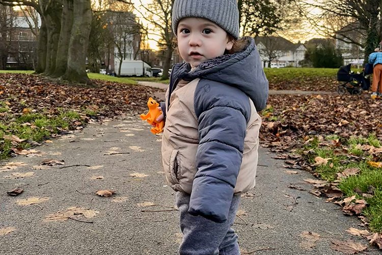 A young boy looks back over his shoulder walking on a path in the park. he is wrapped up warm and holding an orange toy tiger
