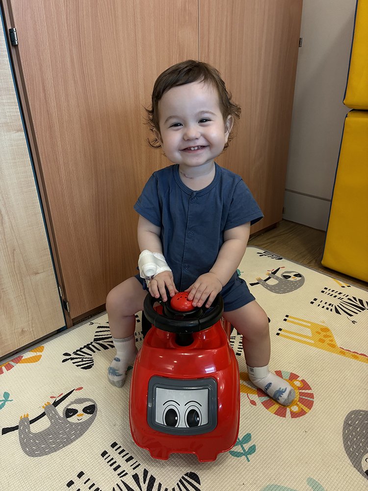 A young boy smiles very broadly at the camera while sitting on a large toy car. He has a medical bandage on his arm.