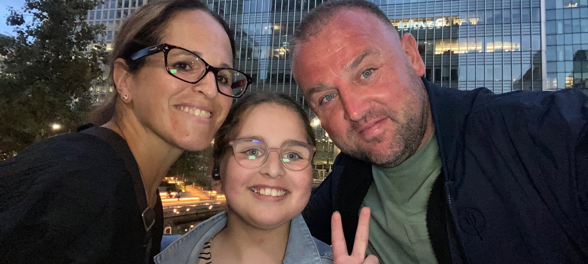 A young girl holds up two of her fingers (the 'peace' sign') with a man and woman leaning in to her, in a selfie pose with a city backdrop. They look happy.