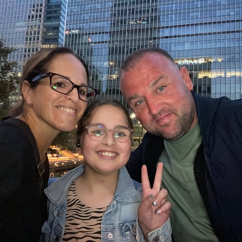 A young girl holds up two of her fingers (the 'peace' sign') with a man and woman leaning in to her, in a selfie pose with a city backdrop. They look happy.