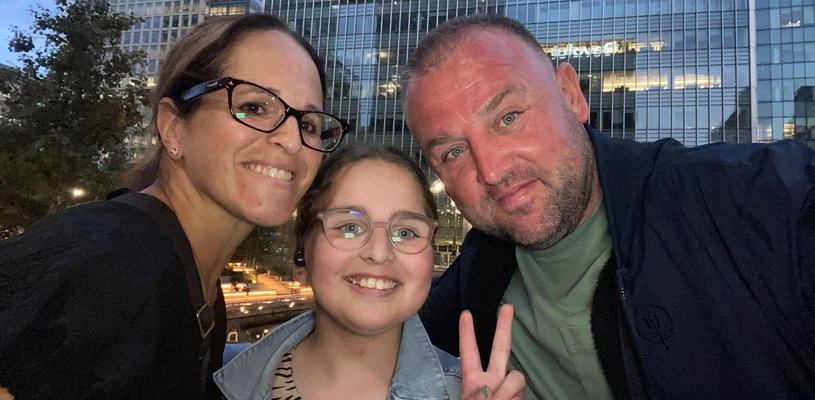 A young girl holds up two of her fingers (the 'peace' sign') with a man and woman leaning in to her, in a selfie pose with a city backdrop. They look happy.