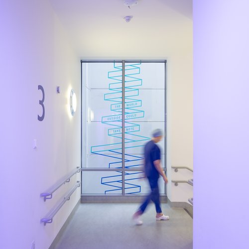 A member of staff in scrubs in the hospital stairwell. There is art work on the window behind them.