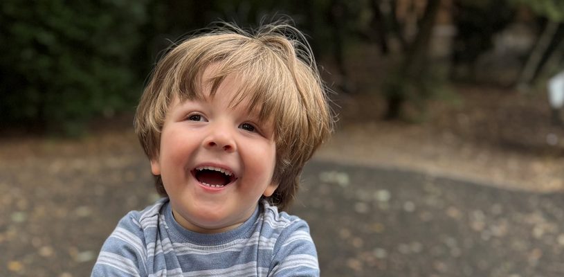 A young boy with brown hair sat on a seesaw. He is smiling and wearing a blue striped top.