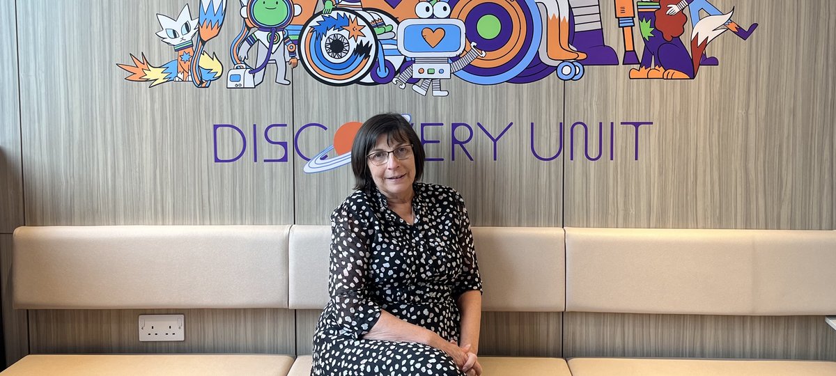 Woman with brown hair sat on white sofa in black and white spotted dress with cartoon space characters in the background