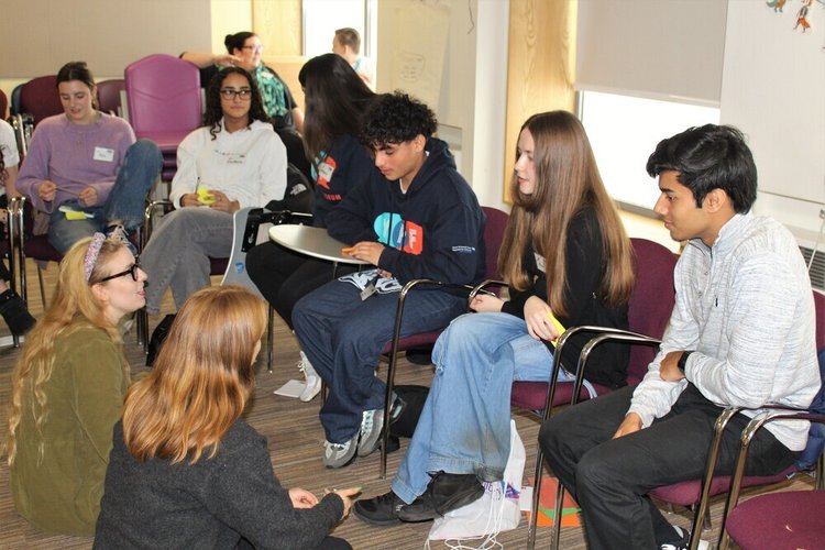A group of teenagers sat in a circle - some on chairs and some sat on the floor - talking.