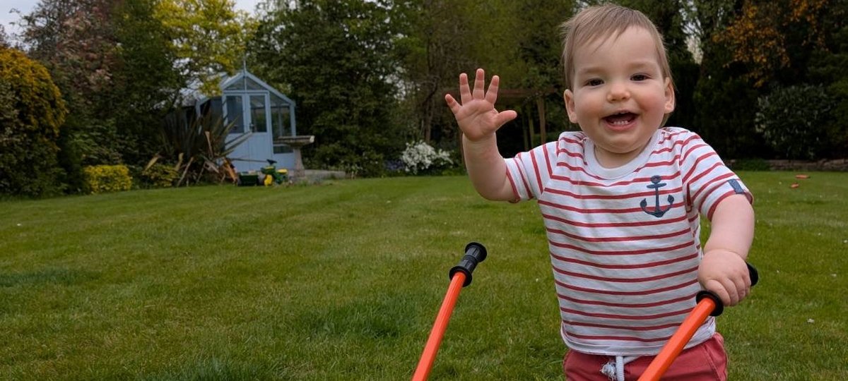Little brown haired boy with a white and red stripped shirt and red shorts waving with a yellow wheel barrow outside
