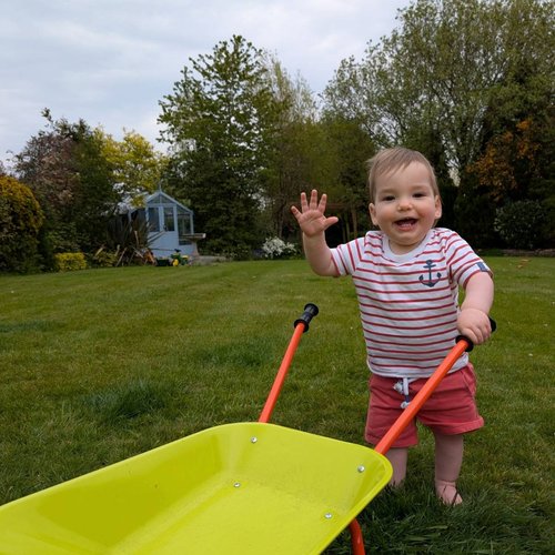 Little brown haired boy with a white and red stripped shirt and red shorts waving with a yellow wheel barrow outside