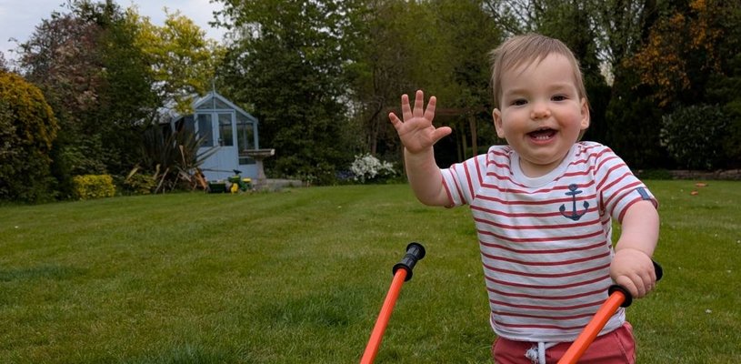 Little brown haired boy with a white and red stripped shirt and red shorts waving with a yellow wheel barrow outside