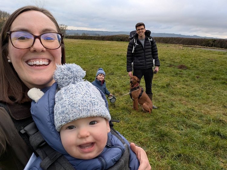 Happy family selfie in a grassy field: smiling woman holds a baby in a knit hat, while a man, child, and dog stand behind them.