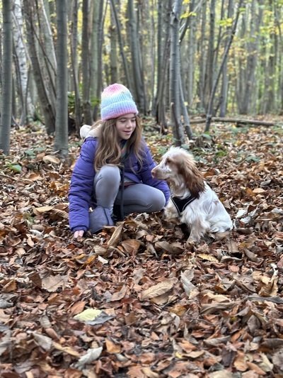 Girl with brown hair in blue jacket with a multi coloured stripped hat sat in a wood surrounded by orange leaves with a brown and white fluffy dog